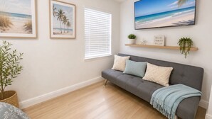 A living room with a grey sofa, two framed beach paintings, a large TV, and a window with blinds. - Peacefull & Central 2-bedroom house with AC in sunny Tampa (Tampa)