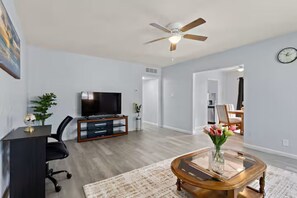 A modern living room with a flat-screen TV, a glass coffee table, a ceiling fan, and a dining area visible through an open doorway. - Casa Serena – Central Tucson Gem w/ Fenced Yard (Tucson)