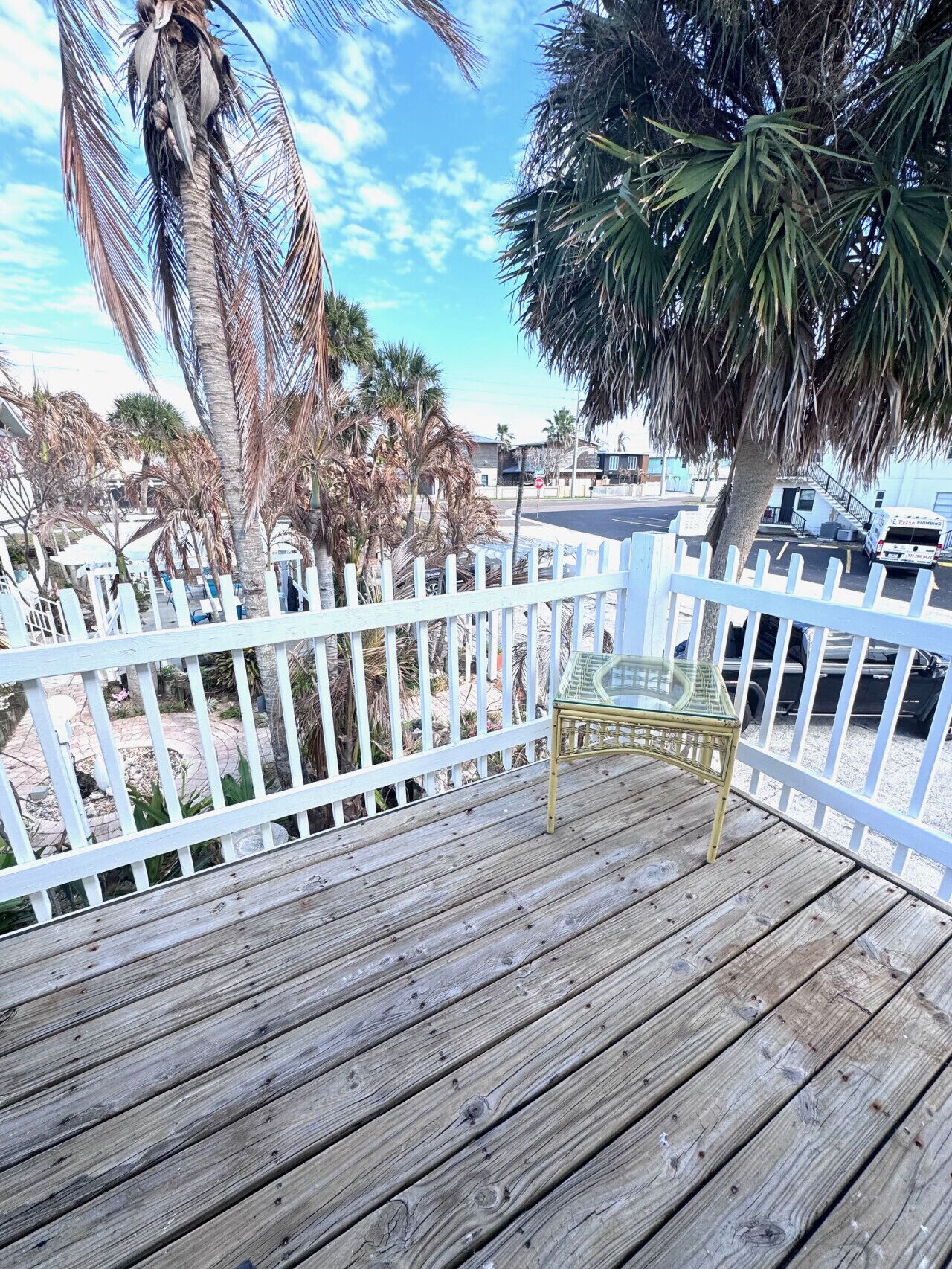 A wooden deck with a glass table and chairs, surrounded by a white fence and palm trees.