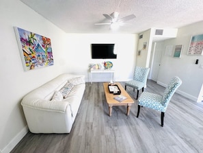 Un salon avec un canapé blanc, deux fauteuils à motifs, une table basse en bois, un ventilateur de plafond et un téléviseur fixé au mur.