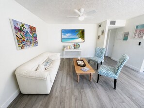 Een woonkamer met een witte bank, een houten salontafel, gestreept stoelen, een plafondventilator en kunstwerken aan de muren.