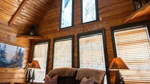 A cozy cabin interior with a sloped ceiling, wooden walls, and large windows with blinds. There is a sofa with cushions and two floor lamps. - Cozy RRG Cabin Hot Tub  Dog Run No-Step Entry (Powell County)