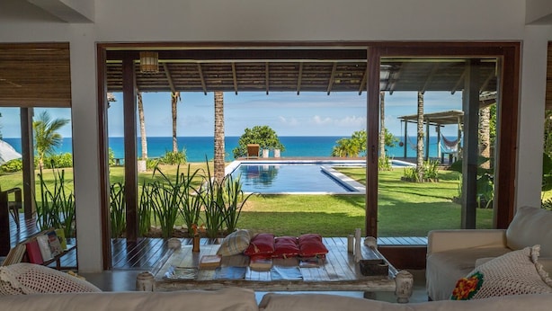 A room with a large window overlooking a pool and the ocean. There is a wooden deck outside the window, and a sofa with cushions inside the room. - Exclusive house in Arraial d'Ajuda: 7 bedrooms, sea view and modern rustic design. (Arraial d'Ajuda)