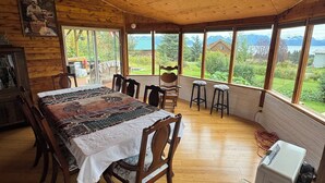 A wooden dining area with a table set for four, a ceiling fan, and a view of mountains through large windows. - Sleeps 4, close to town. Bayviews! (Homer)