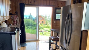 A kitchen with a stainless steel refrigerator, wooden cabinets, a sink, and a window with a view of a house and greenery. - Sleeps 4, close to town. Bayviews! (Homer)
