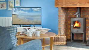 A living room with a TV displaying a beach scene, a fireplace, and a table with cups and a teapot. - Bumblebee Cottage -  a cottage that sleeps 4 guests  in 2 bedrooms (Filey)