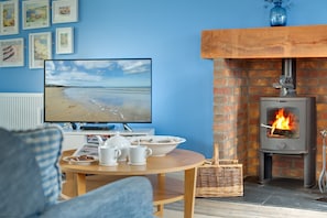A living room with a TV displaying a beach scene, a fireplace, and a table with cups and a teapot. - Bumblebee Cottage - a cottage that sleeps 4 guests in 2 bedrooms (Filey)