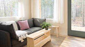 Un salón acogedor con un sofá gris, una pequeña mesa de centro de madera y vistas a un bosque a través de amplios ventanales.