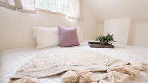Una cama bien hecha con colcha blanca, almohada morada y una manta grande con flecos.