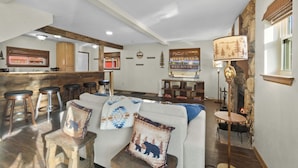 Ein gemütliches Wohnzimmer mit einer Couch, einem Couchtisch und einem Kamin. Im Hintergrund sind Holzschemel und eine Bar zu sehen.