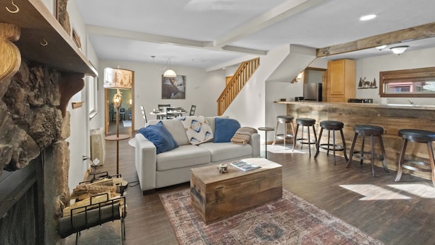 Ein geräumiger Wohnbereich mit einem weißen Sofa, einem Holztisch und einem rustikalen Kamin. Im Hintergrund ist eine Küche mit hölzernen Barhocker und eine Treppe zu sehen.