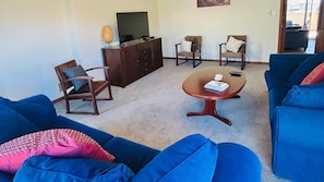 A living room with blue sofas, a wooden coffee table, and two armchairs. A television is mounted on a cabinet, and there is a window providing natural light. - Sea Views Holiday Home- South Bunbury (South Bunbury)