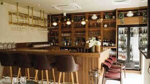 A bar area with a wooden counter, multiple bar stools, shelves with bottles, glasses, and a refrigerator. - The Hillbrook Sherborne (Sherborne)