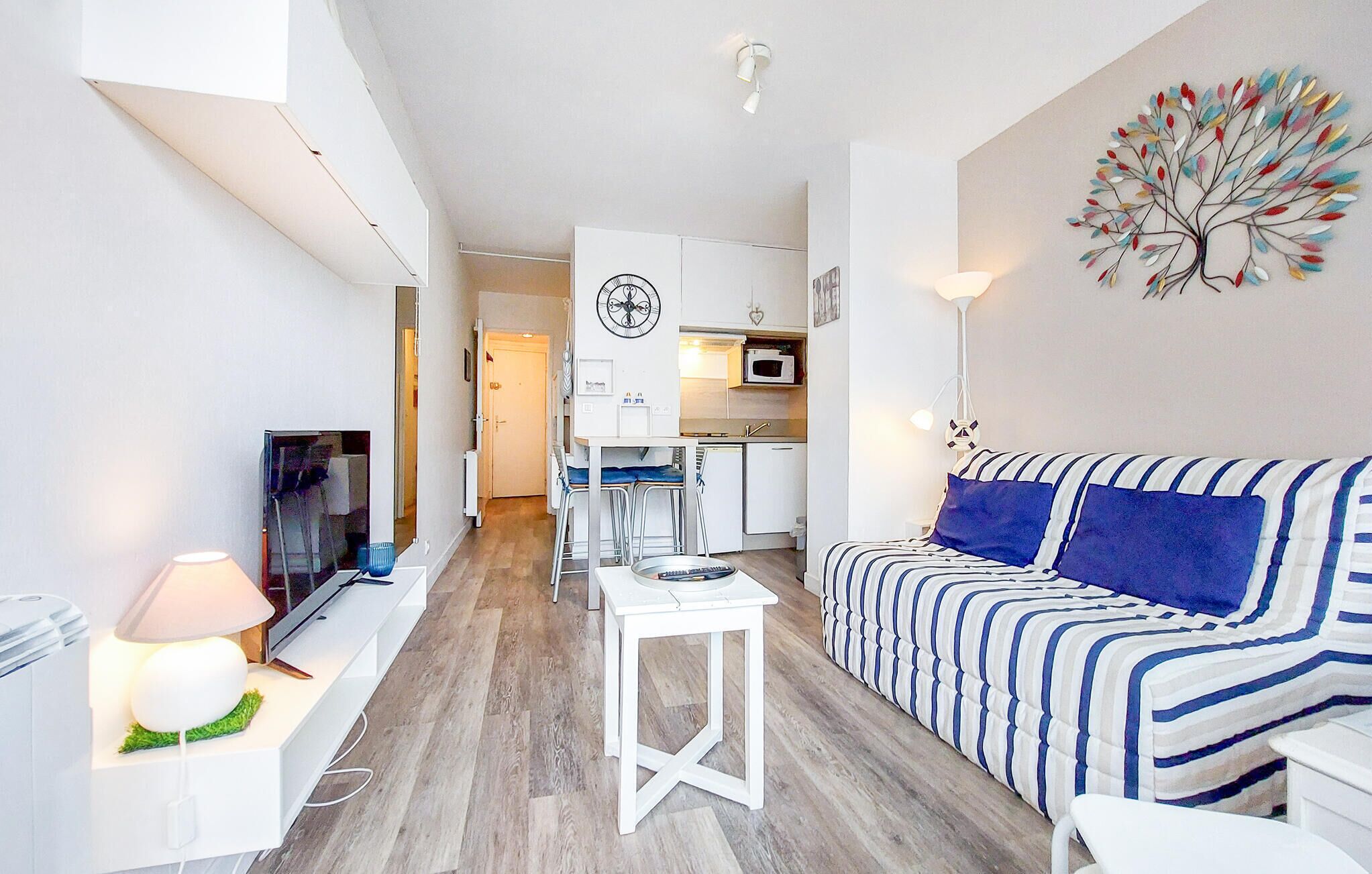 A modern living room with a striped sofa, a small white table, a TV, and a kitchen in the background.