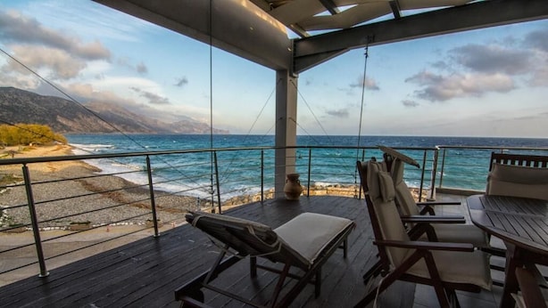 Ein überdachter Außenbereich mit Holzterrasse, Liegestühlen und einem Tisch mit Blick auf Strand und Meer.