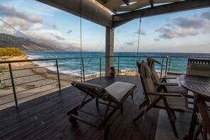A covered outdoor area with a wooden deck, lounge chairs, and a table overlooking a beach and ocean. - Gonis Suites - Paleochora (Kantanos-Selino)