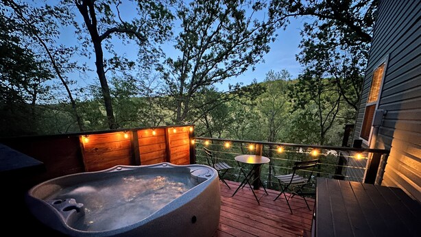 A hot tub on a wooden deck with string lights, surrounded by trees. - Lakefront w/ hot tub, firepit, BBQ, smoker, kitchen, patios, king bed, 1.5 bath (Eureka Springs)
