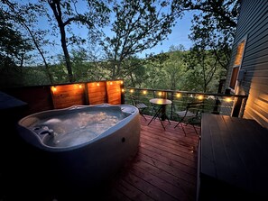 A hot tub on a wooden deck with string lights, surrounded by trees. - Lakefront w/ hot tub, firepit, BBQ, smoker, kitchen, patios, king bed, 1.5 bath (Eureka Springs)