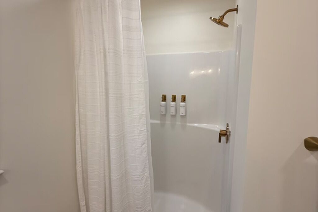 A shower area with a glass door, a white shower curtain, and three bottles of shampoo on the wall.