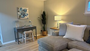 Ett vardagsrum med en grå soffa, ett litet skrivbord med en stol, en stor växt och en lampa.