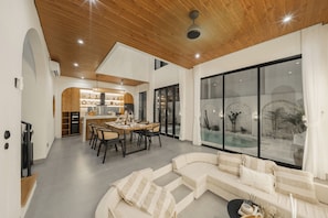 A modern living room with a sectional sofa, a dining area with a wooden table, and a large glass window overlooking a pool. - Villa Juana  (Canggu)