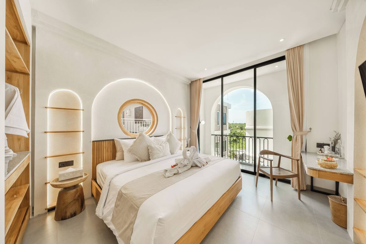 Quarto de hotel moderno com uma cama grande, um espelho redondo, uma varanda com uma cadeira e uma mesinha de madeira.
