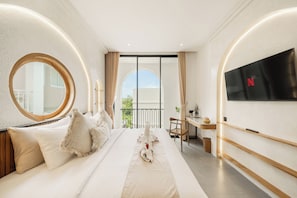 A hotel room with a large bed, a round mirror, a desk, a TV, and a balcony view. - Villa Juana  (Canggu)