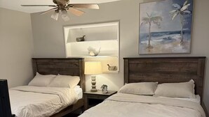 A bedroom with two wooden beds, a ceiling fan, a shelf with decorative items, and a painting of palm trees. - Spa Beach Retreat • Sleeps 10 • 8 min to Beach (Englewood)