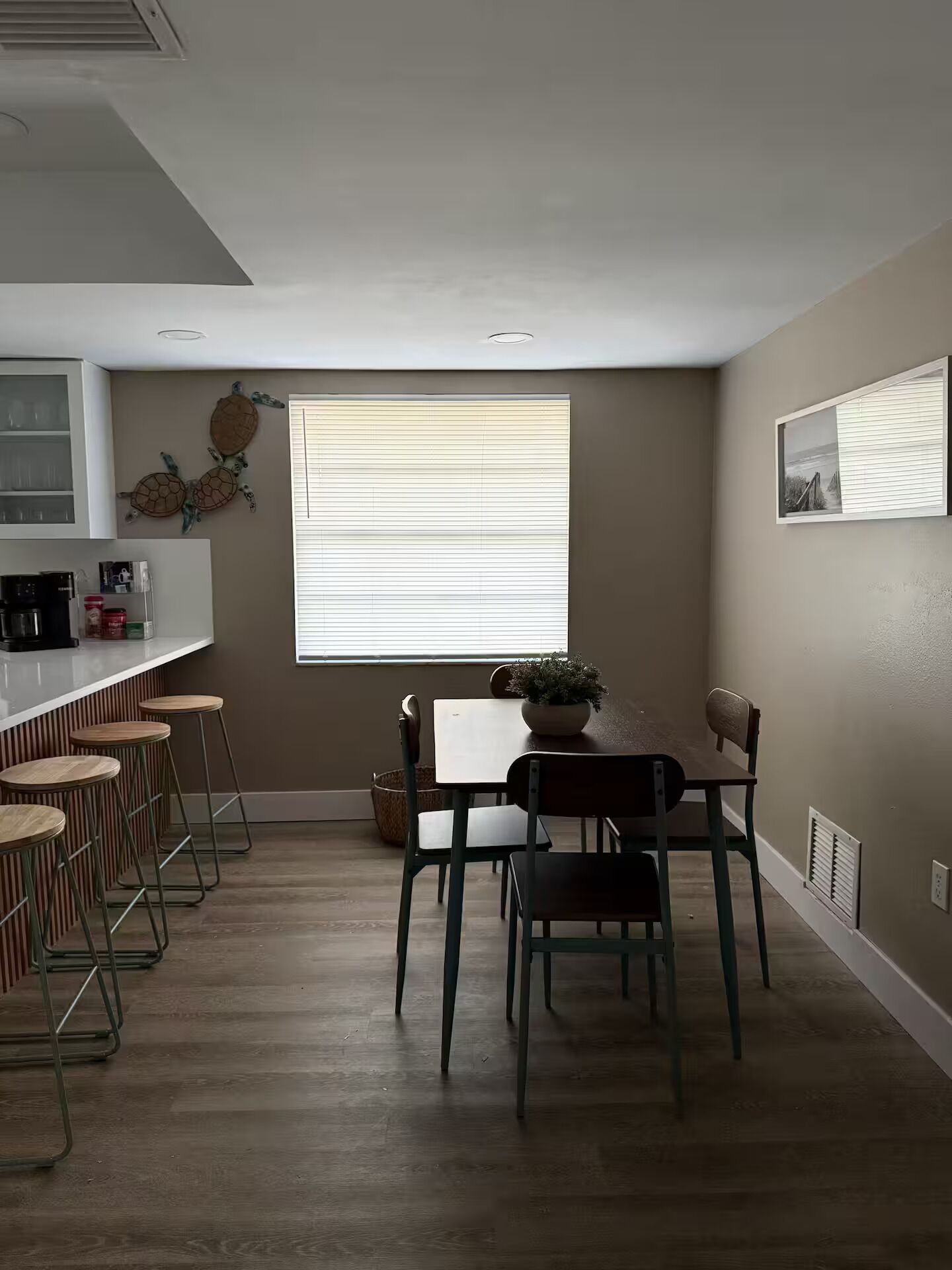 A dining area with a table and chairs, a window with blinds, a kitchen counter with stools, and a wall-mounted shelf.