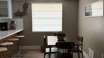 A dining area with a table and chairs, a window with blinds, a kitchen counter with stools, and a wall-mounted shelf.