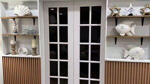 A room with a wooden floor, white walls, and a white door with glass panels. Shelves on the walls hold decorative items like seashells, fish, and candles. - Spa Beach Retreat • Sleeps 10 • 8 min to Beach (Englewood)