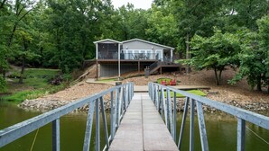 A house with a deck and a dock leading to a pond. - Spacious Lakefront Getaway with Dock in Quiet Cove (Lake Ozark)