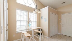 Una habitación con zona de comedor, una mesa blanca y bancos, una ventana con persianas y una puerta.