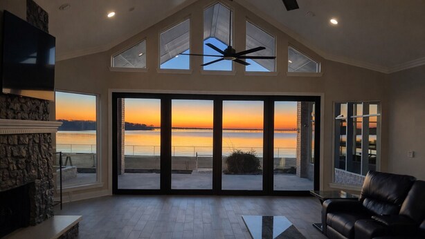 A spacious living room with a large sliding glass door leading to a balcony overlooking a body of water at sunset. - Spacious 6-bedroom house on lake with extra large kitchen and great swimming. (Corsicana)