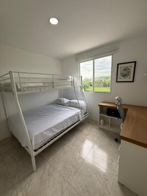 A single-bed room with a bunk bed, a small desk, a chair, a window with a view of greenery, and a ceiling light.