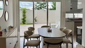 Eine moderne Küche mit weißen Schränken, ein Essbereich mit einem runden Tisch und sechs Stühlen sowie ein Blick auf einen Garten durch große Schiebeglastüren.