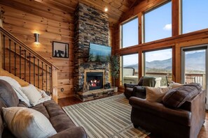 A cozy living room with a stone fireplace, leather sofas, a large window, and a wooden staircase. - VIEWS/Hot Tub/Fire Pit/Game Room/Daybed Swing - Three Bears Chalet (Gatlinburg)