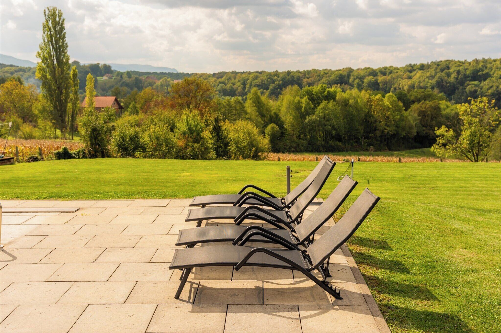 A row of black lounge chairs on a paved patio with a grassy lawn and trees in the background.