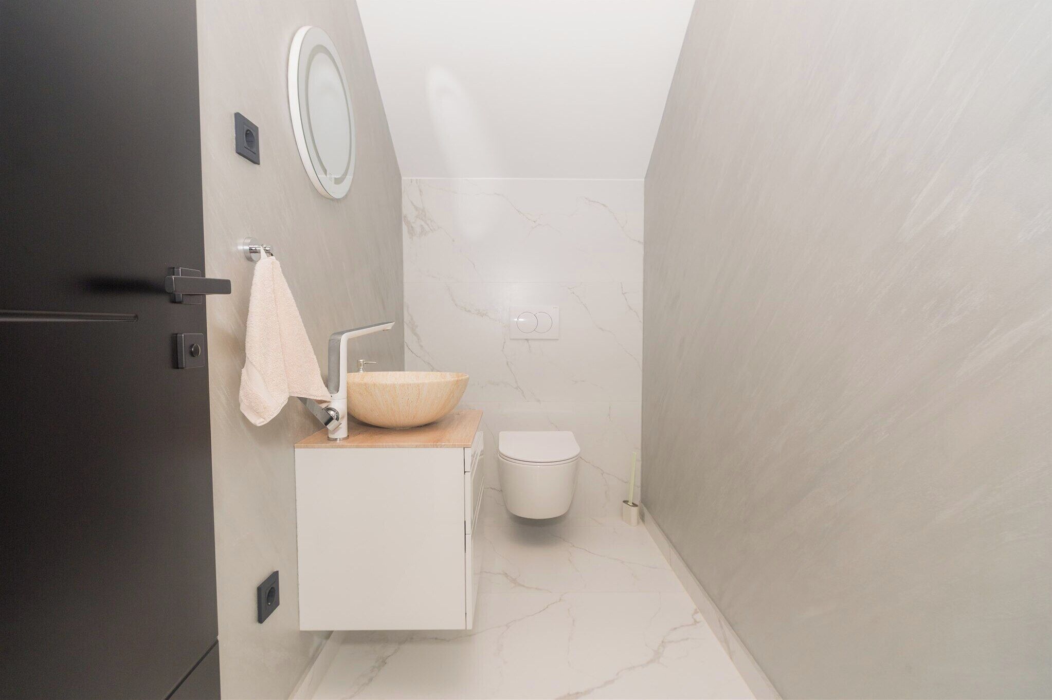 A compact bathroom with a wooden vanity, a round mirror, a white toilet, and a beige towel hanging on the door.