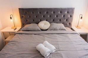 Un letto rifatto con cura, con testiera trapuntata, due abat-jour e una coppia di asciugamani arrotolati sul letto.