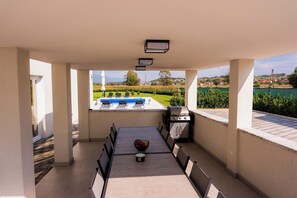 Un'area esterna coperta con tavolo e sedie, un barbecue e vista su una piscina e del verde.