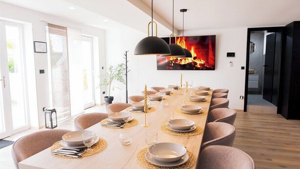 Un comedor moderno con una mesa larga dispuesta para una comida, luces colgantes y una chimenea al fondo.