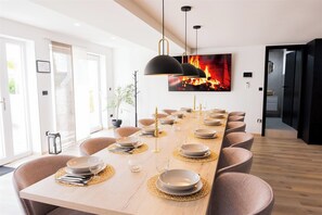 Uma sala de jantar moderna com uma mesa comprida posta para uma refeição, luminárias pendentes e uma lareira ao fundo.