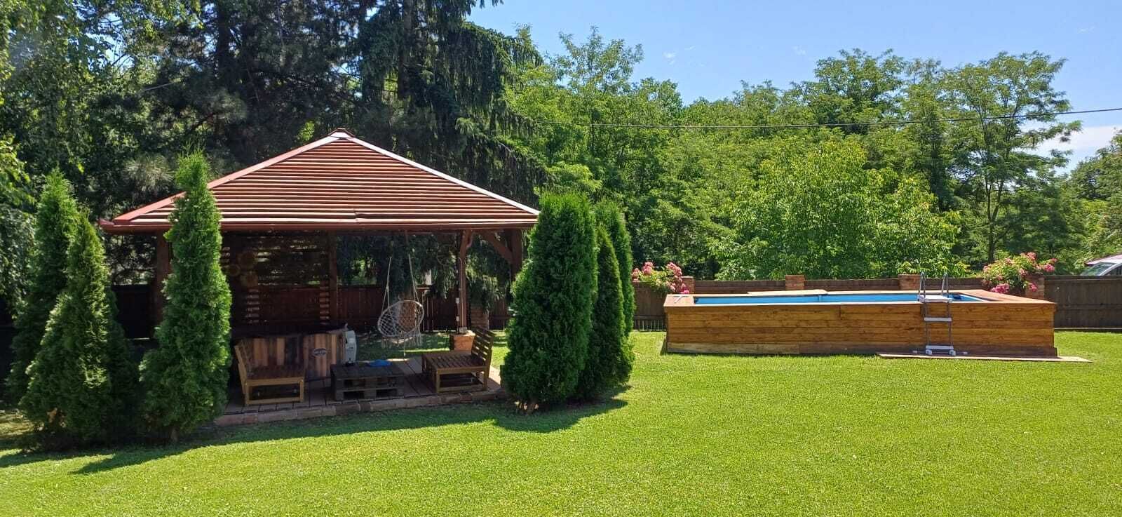 A wooden gazebo with a sloped roof, surrounded by a well-maintained lawn and tall trees.