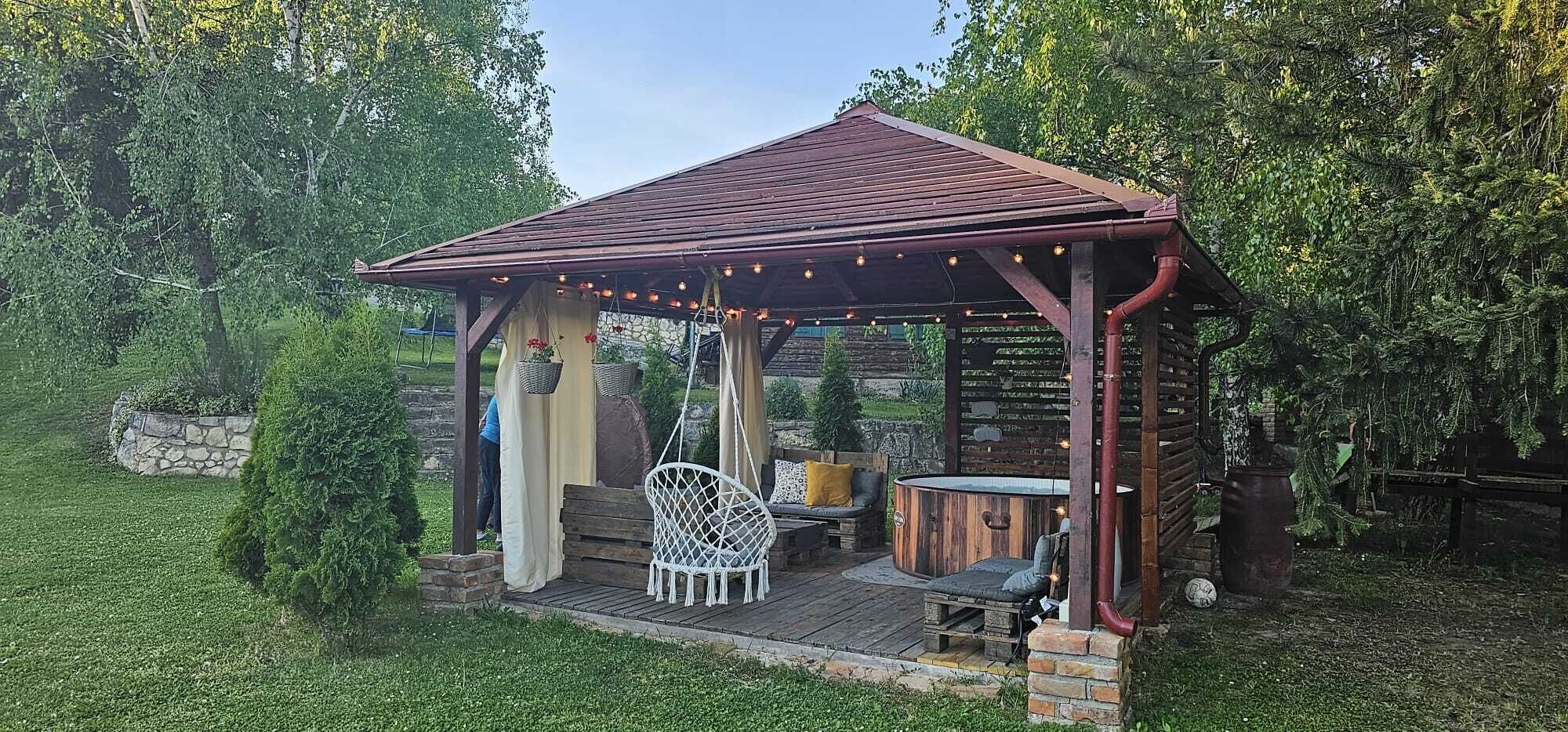 A wooden gazebo with a sloped roof, a hanging chair, a bench, and a small table with a stool.