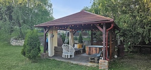 A wooden gazebo with a sloped roof, a hanging chair, a bench, and a small table with a stool.