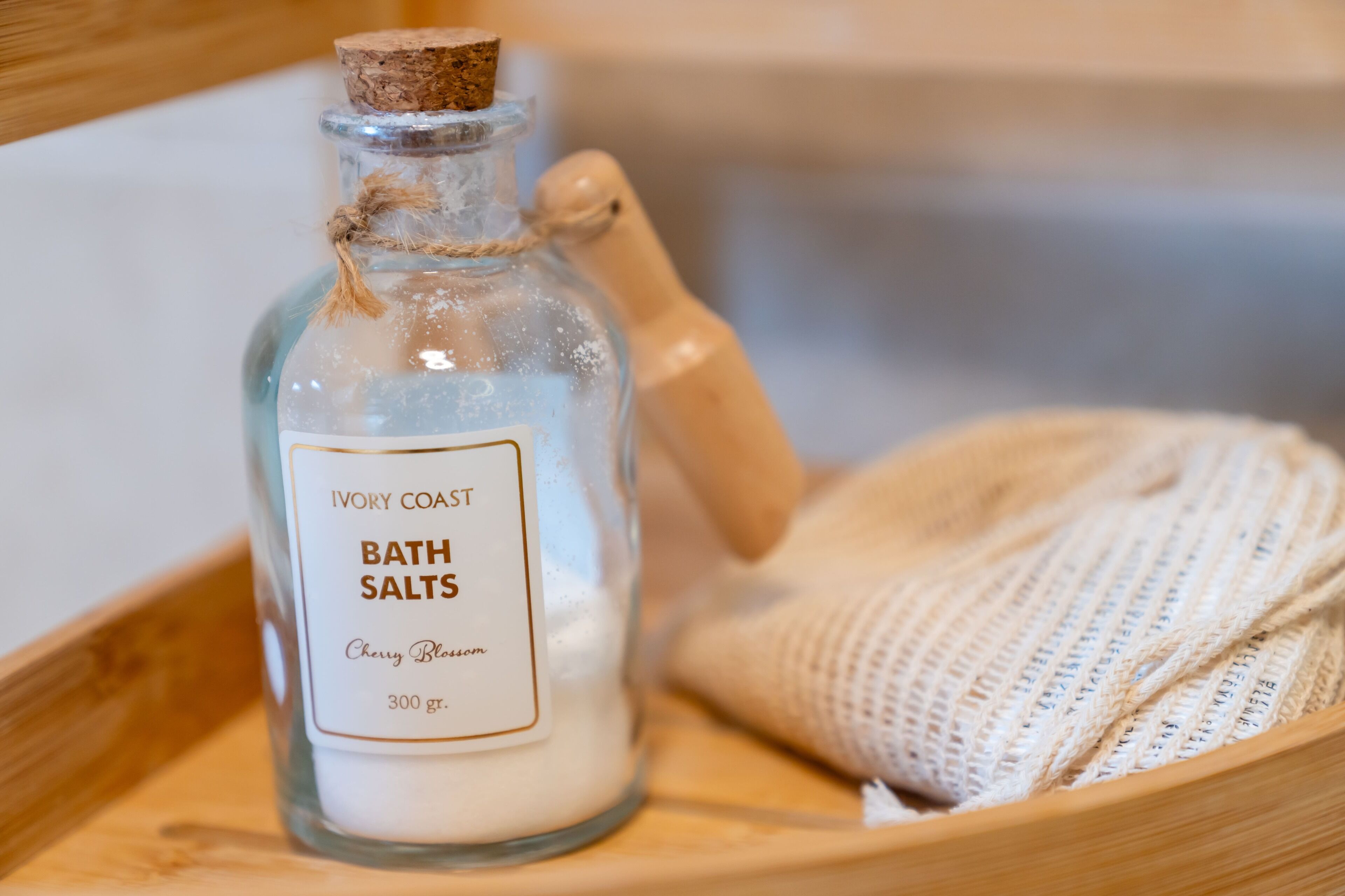 Una botella de vidrio etiquetada como "Ivory Coast Bath Salts", con un cucharón de madera y una tela tejida en una bandeja de madera.