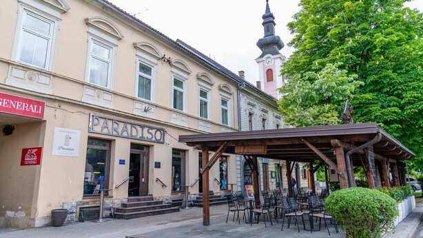 Un immeuble avec une enseigne indiquant « Appartements Paradiso », un espace extérieur abrité avec des sièges, et un clocher d’église en arrière-plan.