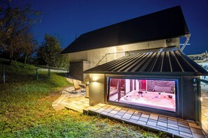 Ein modernes Haus mit einem durch Glas geschützten Whirlpoolbereich, einer Terrasse mit Grill und einer Treppe, die zum oberen Stockwerk führt.