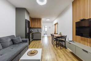 A modern apartment with a grey sofa, a dining area with a wooden table and chairs, a kitchen with grey cabinets, and a flat-screen TV mounted on the wall. - City Center Apartment Cozy 1BR With Patio (Tirana)